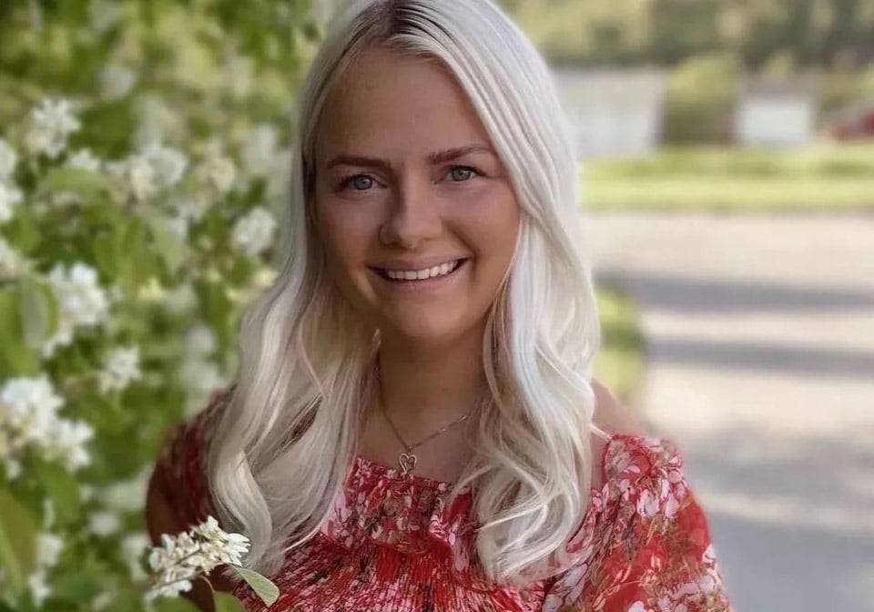 Eine blonde Frau mit sonnengeküsster Haut lächelt vor einem grünen Hintergrund.