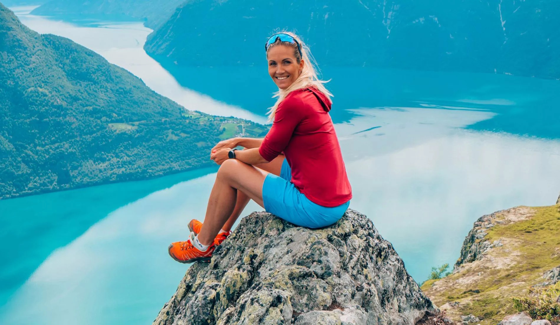 Eine gebräunte Frau sitzt auf einem Felsen in der Nähe einer Berglandschaft.